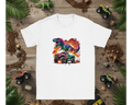 White t-shirt with colorful dinosaur and monster truck graphic on a wooden background with toy trucks and leaves.
