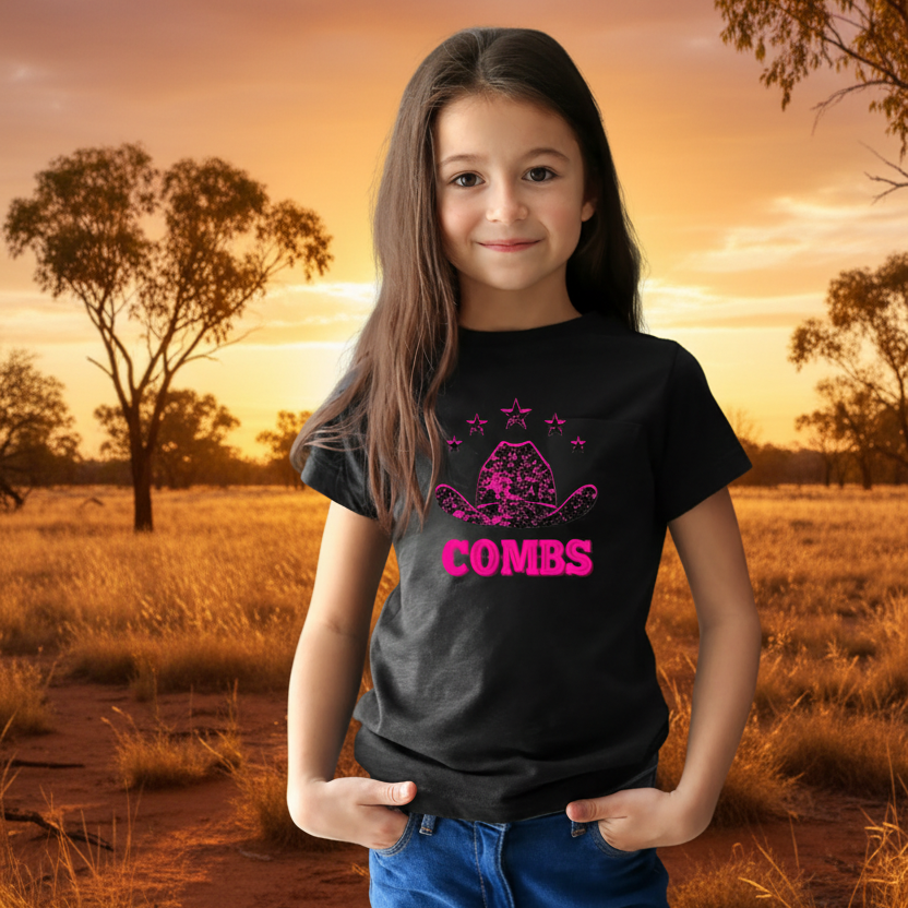Young girl wearing a black t-shirt with pink graphic design and text, standing against a dark brick wall.