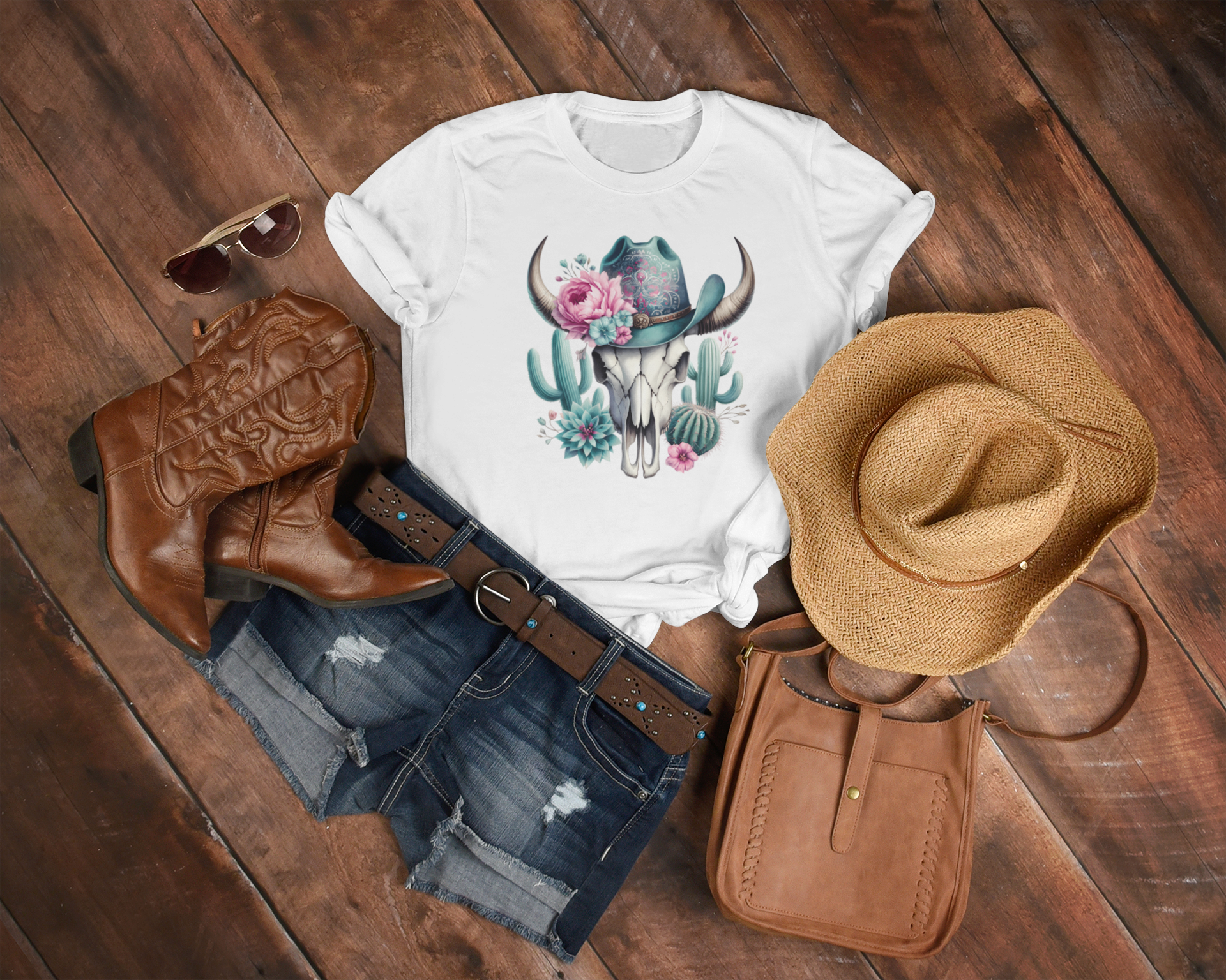 White t-shirt with a graphic design, cowboy boots, denim shorts, hat, and purse on a wooden surface