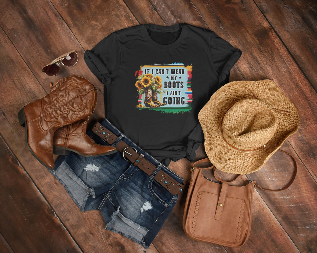 Black t-shirt with a colorful graphic and text, paired with cowboy boots, shorts, a hat, and a bag on a wooden surface.